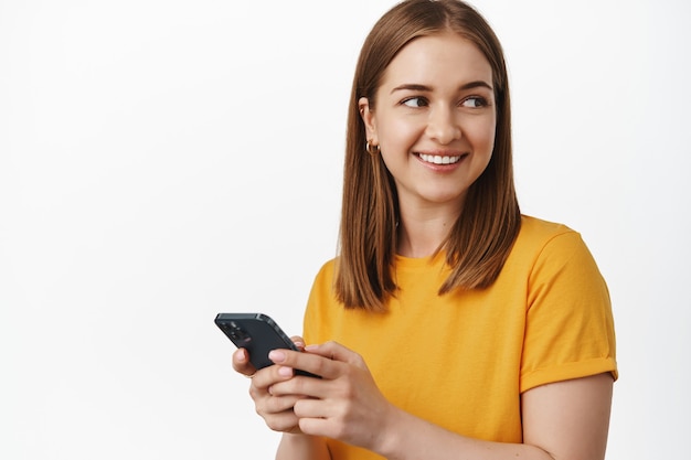 Glimlachende jonge vrouw gebruikt smartphone, houdt mobiele telefoon app vast, draait hoofd achter schouder met blij gezicht, staande in geel t-shirt tegen witte muur