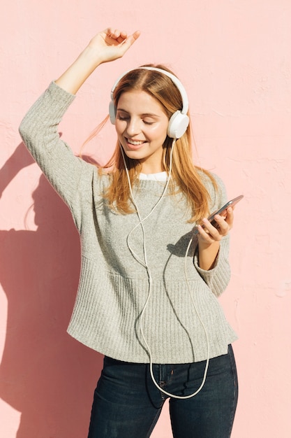 Glimlachende jonge vrouw het luisteren muziek op hoofdtelefoon die tegen roze muur dansen