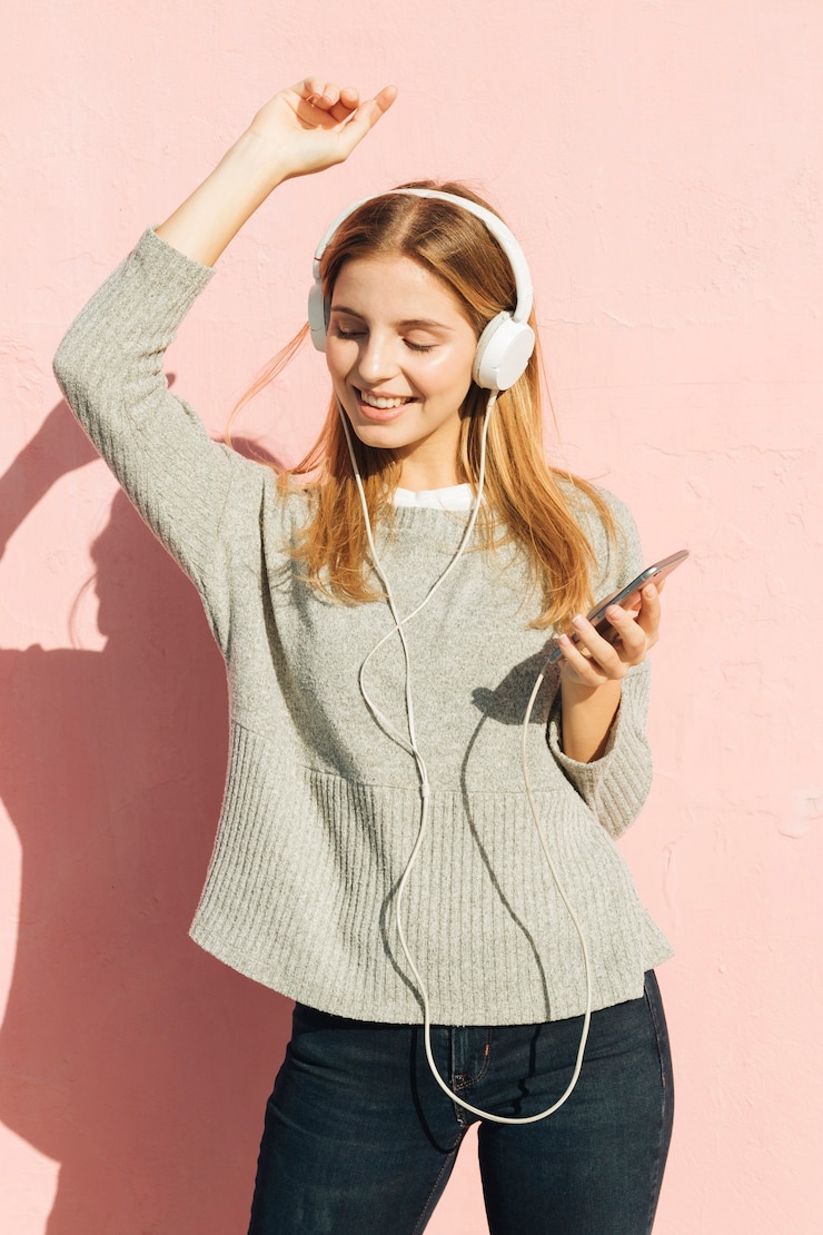 Gratis Foto glimlachende jonge vrouw het luisteren muziek op hoofdtelefoon die tegen roze muur dansen