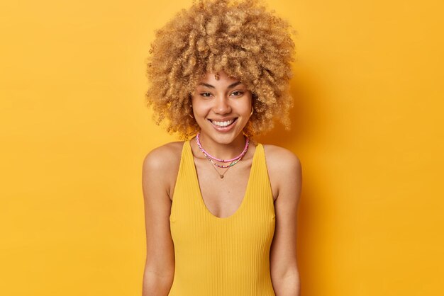 Glimlachende jonge vrouw met krullend borstelig haar gekleed in casual t-shirt heeft blije uitdrukking drukt positieve emoties uit geïsoleerd over levendige gele achtergrond hoort goed nieuws Mensen en geluk