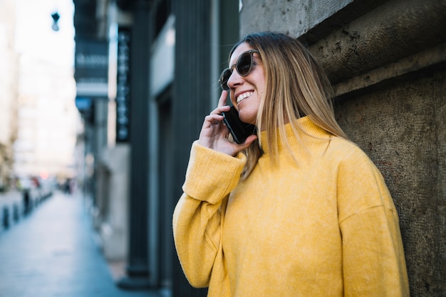 Gratis foto glimlachende jonge vrouw met zonnebril en smartphone die dichtbij op straat voortbouwen