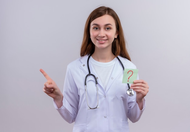 Glimlachende jonge vrouwelijke arts die medisch gewaad en stethoscoop draagt die document nota met vraagteken op het geschreven houdt en naar de linkerkant op geïsoleerde witte muur richt