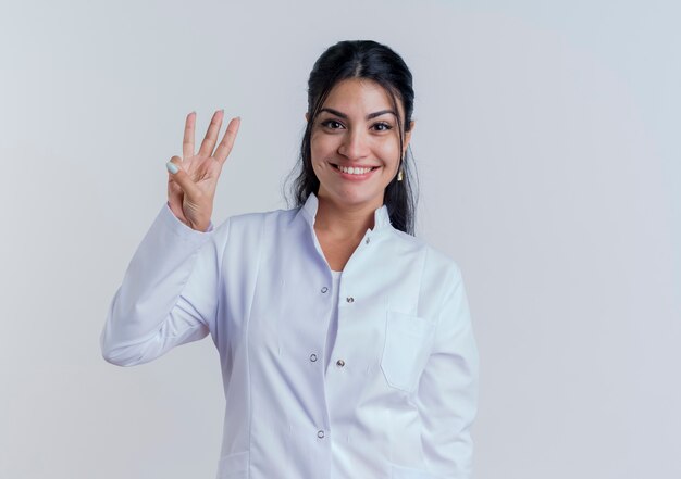 Glimlachende jonge vrouwelijke arts die medisch kleed draagt dat en drie met geïsoleerde hand kijkt toont
