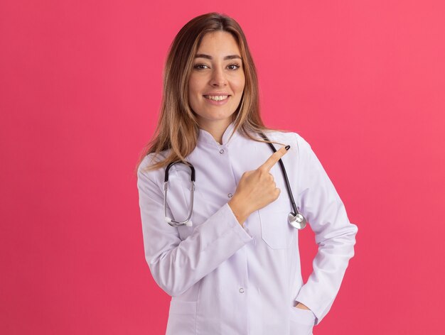 Glimlachende jonge vrouwelijke arts die medisch kleed met stethoscooppunten aan kant draagt die op roze muur met exemplaarruimte wordt geïsoleerd