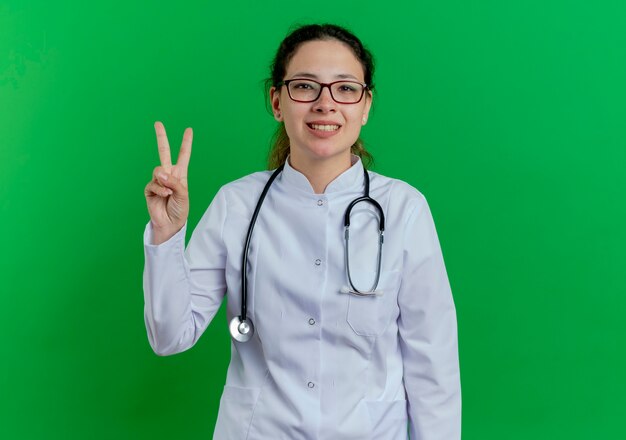Glimlachende jonge vrouwelijke arts die medische mantel en stethoscoop en bril draagt die vredesteken doet dat op groene muur met exemplaarruimte wordt geïsoleerd