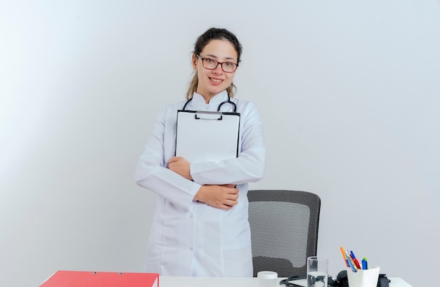 Glimlachende jonge vrouwelijke arts die medische mantel en stethoscoop en glazen draagt die zich achter bureau met medische hulpmiddelen bevinden die geïsoleerd holdingsklembord kijken