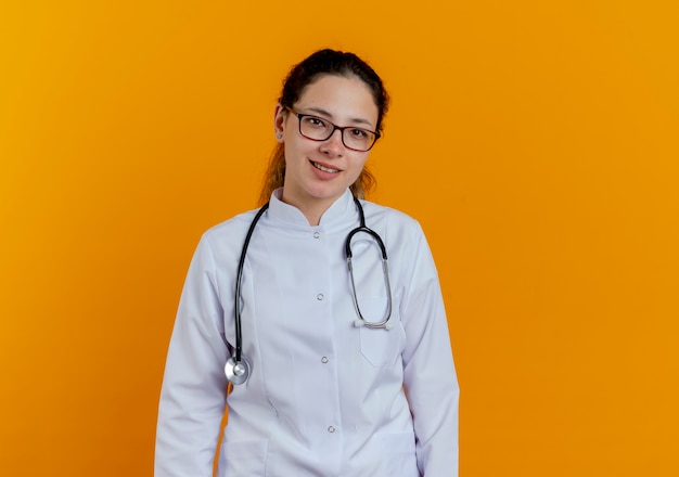 Glimlachende jonge vrouwelijke arts die medische mantel en stethoscoop met geïsoleerde bril draagt