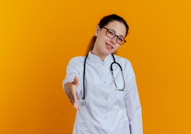 Glimlachende jonge vrouwelijke arts die medische mantel en stethoscoop met glazen die geïsoleerde hand standhouden