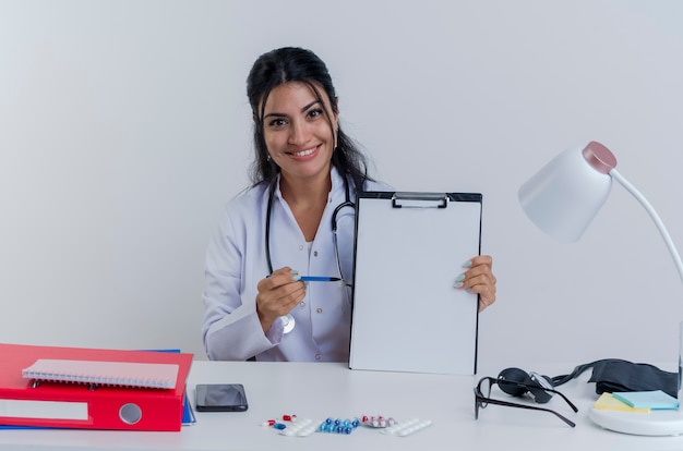 Glimlachende jonge vrouwelijke arts die medische mantel en stethoscoopzitting bij bureau met medische hulpmiddelen draagt die het tonen van klembord die met pen richten op geïsoleerde het kijken