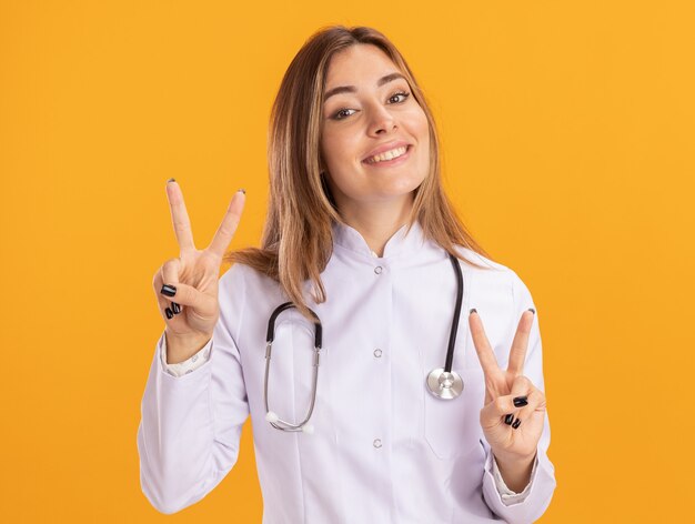 Glimlachende jonge vrouwelijke arts die medische mantel met stethoscoop draagt die vredesgebaar toont dat op gele muur wordt geïsoleerd