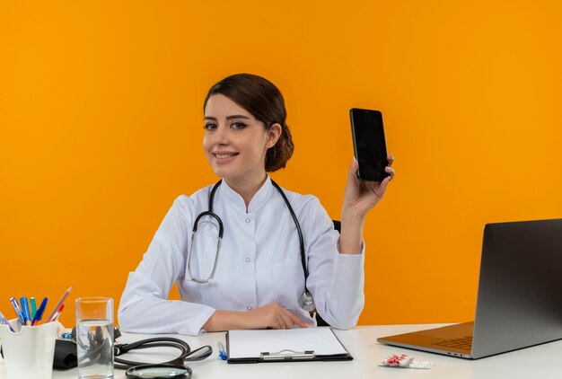 Glimlachende jonge vrouwelijke arts die medische mantel met stethoscoop zittend aan bureau werkt op computer met medische hulpmiddelen die telefoon op isolatie gele achtergrond houden