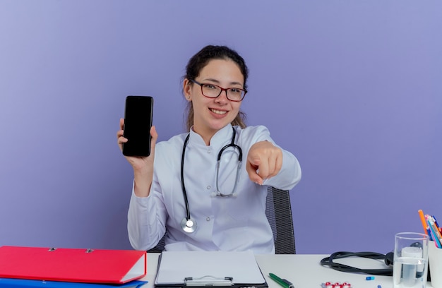 Glimlachende jonge vrouwelijke arts die medische robe en stethoscoopzitting bij bureau met medische hulpmiddelen draagt die mobiele telefoon toont