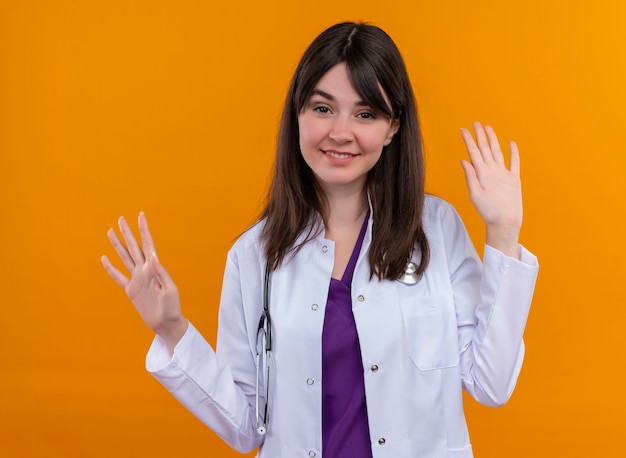 Glimlachende jonge vrouwelijke arts in medische mantel met stethoscoop werpt handen omhoog op geïsoleerde witte achtergrond oranje exemplaarruimte