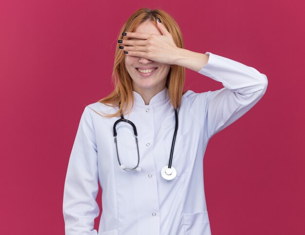 Glimlachende jonge vrouwelijke gemberdokter die een medische mantel en een stethoscoop draagt die de ogen bedekt met de hand