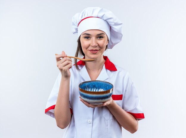 Glimlachende jonge vrouwelijke kok die een uniforme chef-kok draagt die kom met lepel draagt die op witte muur wordt geïsoleerd