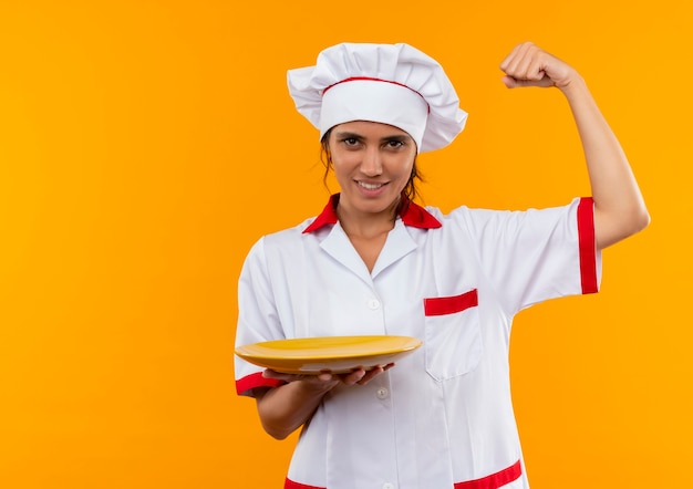 Glimlachende jonge vrouwelijke kok die plaat van de chef-kok de eenvormige holding draagt en sterk gebaar met exemplaarruimte toont