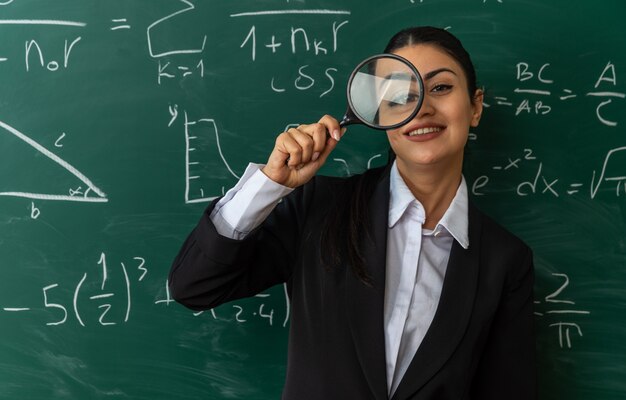 Glimlachende jonge vrouwelijke leraar die voor het schoolbord staat en naar de camera kijkt met vergrootglas in de klas