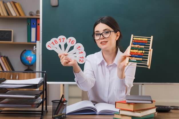 glimlachende jonge vrouwelijke leraar met een bril die een telraam vasthoudt met een nummerventilator die aan een bureau zit met schoolhulpmiddelen in de klas