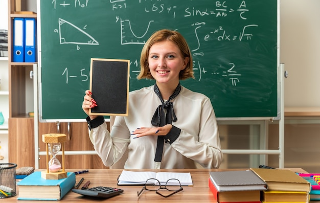 glimlachende jonge vrouwelijke leraar zit aan tafel met schoolbenodigdheden en wijst met de hand naar mini schoolbord in de klas