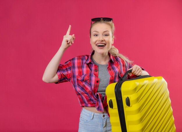 Glimlachende jonge vrouwelijke reiziger die rood overhemd in glazen draagt die koffer wijst op geïsoleerde roze muur