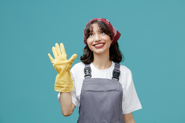 Glimlachende jonge vrouwelijke schoonmaker die uniforme bandana en rubberen handschoenen draagt en omhoog kijkt met hallo gebaar geïsoleerd op blauwe achtergrond