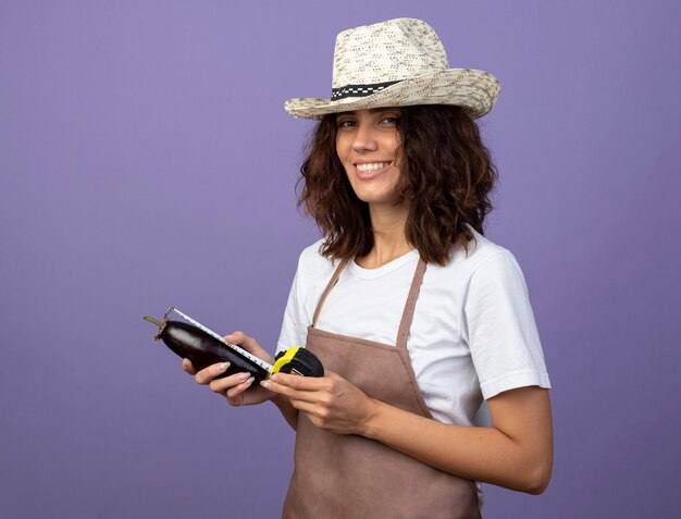Glimlachende jonge vrouwelijke tuinman in eenvormig die het tuinieren hoed draagt die aubergine met meetlint meet