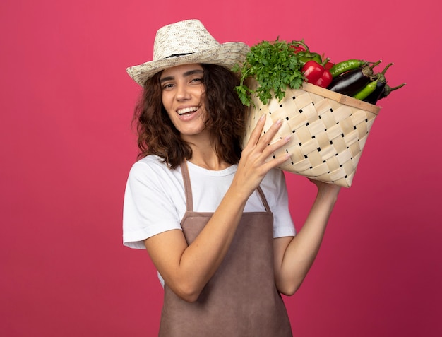 Glimlachende jonge vrouwelijke tuinman in uniform die tuinieren hoed draagt die plantaardige mand op schouder houdt die op roze wordt geïsoleerd