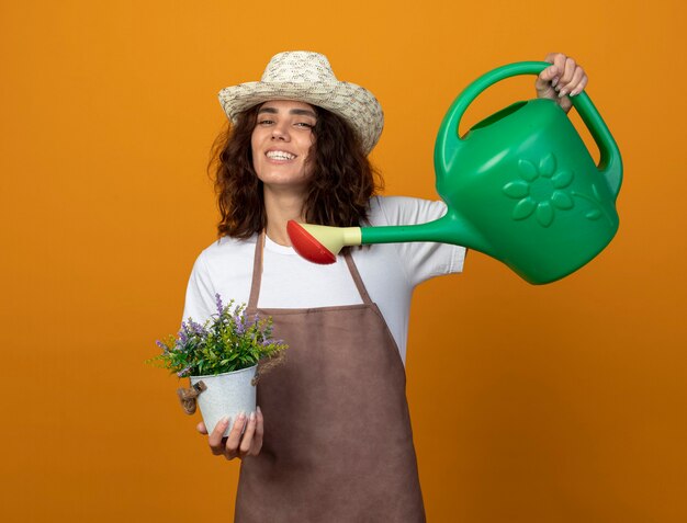 Glimlachende jonge vrouwelijke tuinman in uniform dragen tuinieren hoed drenken bloem in bloempot met gieter geïsoleerd op oranje muur