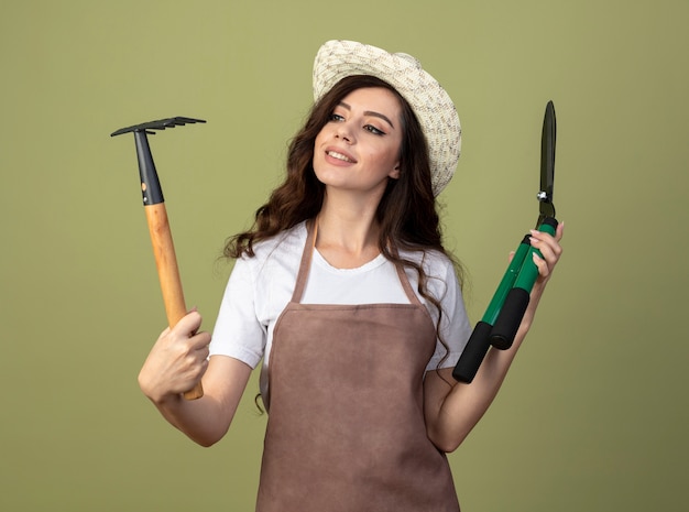 Glimlachende jonge vrouwelijke tuinman in uniform dragen tuinieren hoed houdt tondeuse en kijkt naar hark geïsoleerd op olijfgroene muur
