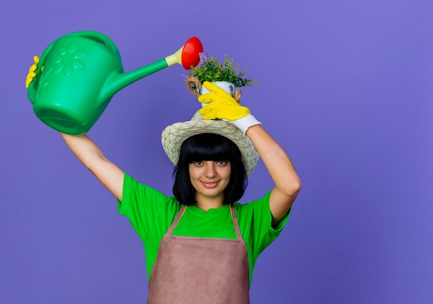 Glimlachende jonge vrouwelijke tuinman in uniform met tuinhoed doet alsof ze bloemen water geeft