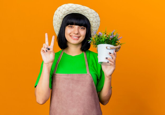 Glimlachende jonge vrouwelijke tuinman in uniform met tuinhoed houdt bloempot vast