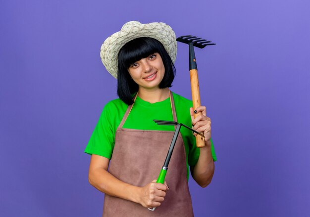 Glimlachende jonge vrouwelijke tuinman in uniform met tuinhoed houdt hark en schoffelhark vast