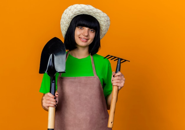 Glimlachende jonge vrouwelijke tuinman in uniform met tuinhoed houdt schop en hark vast