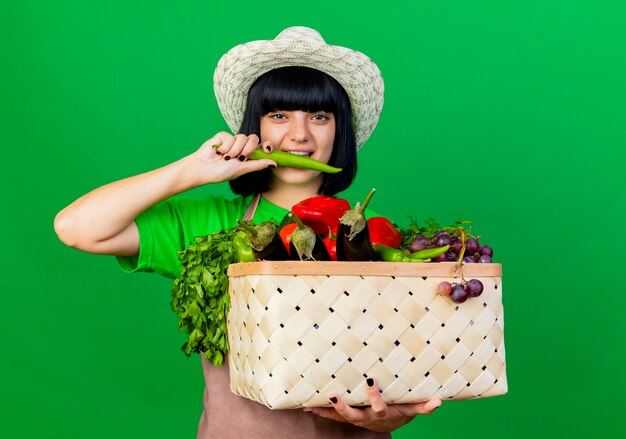 Glimlachende jonge vrouwelijke tuinman in uniform met tuinhoed met groentemand