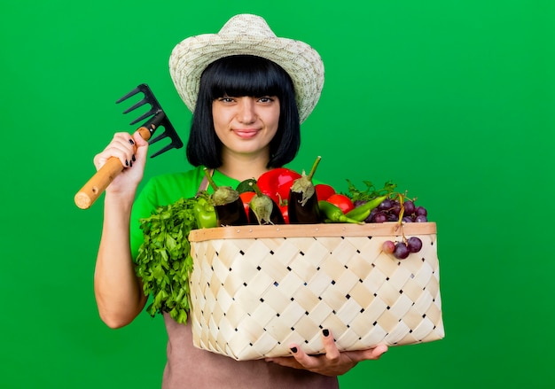 Glimlachende jonge vrouwelijke tuinman in uniform met tuinhoed met groentemand