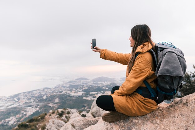 Glimlachende jonge vrouwelijke wandelaarzitting bovenop berg die selfie op mobiele telefoon nemen