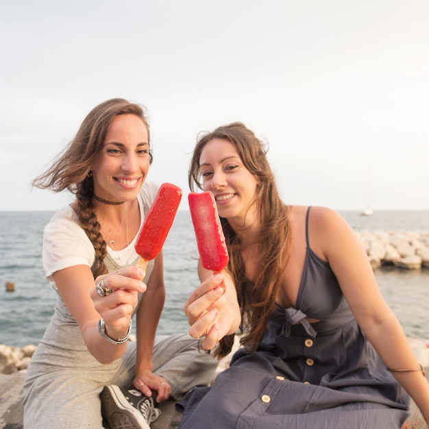 Glimlachende jonge vrouwen die op kust zitten die rode ijslollys tonen