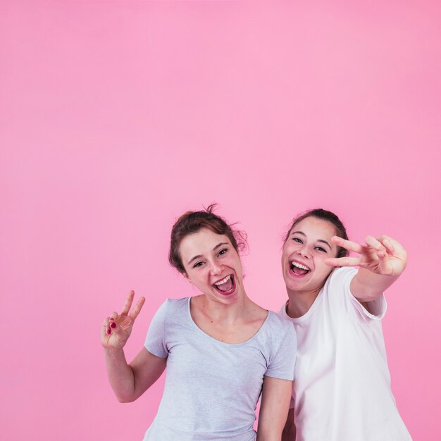 Glimlachende jonge vrouwen die vredesteken tonen die zich tegen roze achtergrond bevinden