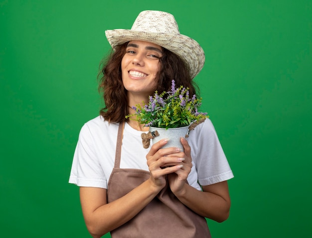 Gratis foto glimlachende jonge vrouwentuinman in uniform die het tuinieren de bloem van de hoedenholding in bloempot dragen die op groen wordt geïsoleerd