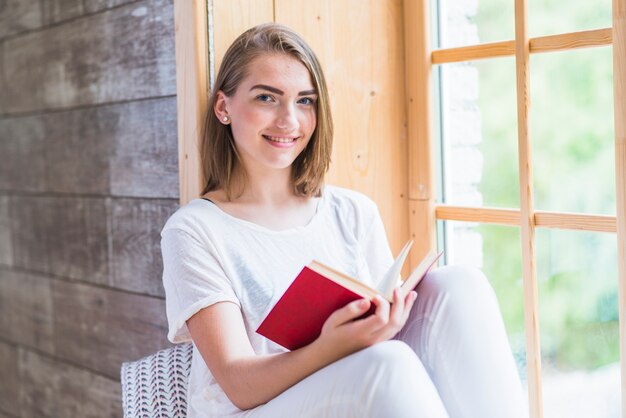 Glimlachende jonge vrouwenzitting dichtbij het boek van de vensterlezing
