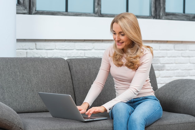 Glimlachende jonge vrouwenzitting op bank die laptop thuis met behulp van