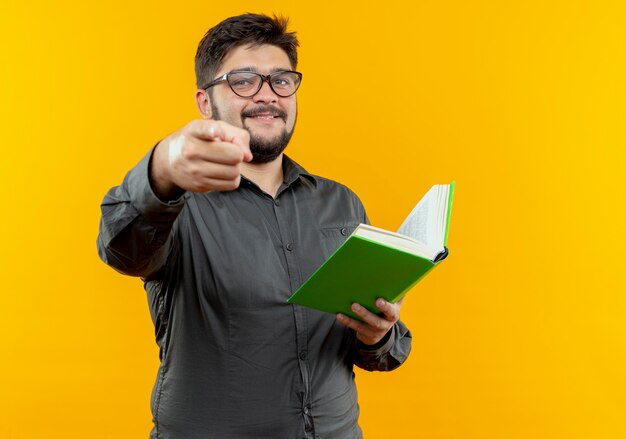 Glimlachende jonge zakenman die een bril draagt die boek houdt en u gebaar toont dat op gele muur wordt geïsoleerd