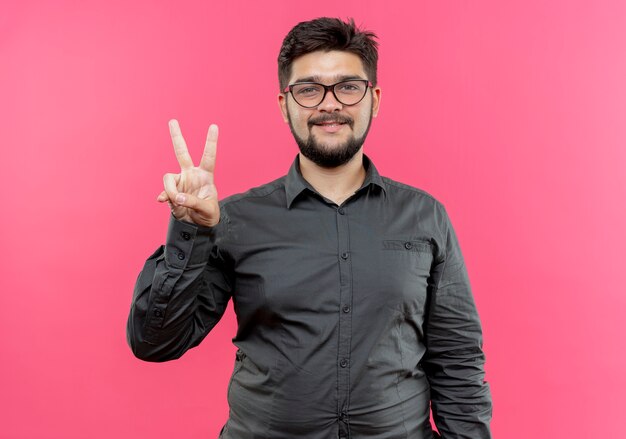 Glimlachende jonge zakenman die glazen draagt die vredesgebaar tonen dat op roze wordt geïsoleerd