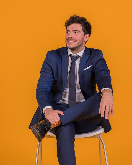 Glimlachende jonge zakenmanzitting op stoel die weg tegen een oranje achtergrond kijken