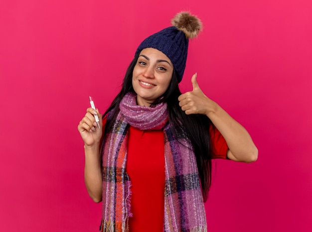 Glimlachende jonge zieke vrouw die de winterhoed en de thermometer van de sjaalholding voorzijde bekijkt die duim toont die omhoog op roze muur met exemplaarruimte wordt geïsoleerd
