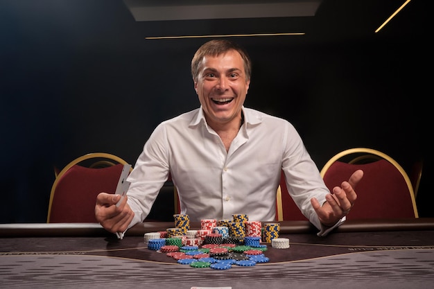 Glimlachende jongeman in een wit klassiek overhemd speelt poker aan de tafel in het casino. Hij verheugt zich in het winnen en kijken naar de camera. Gokken voor geld. Spelen van fortuin.