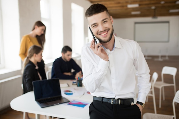 Glimlachende jongeman met baard in wit overhemd die vreugdevol in de camera kijkt terwijl hij op mobiel praat terwijl hij tijd doorbrengt op kantoor met collega's op de achtergrond