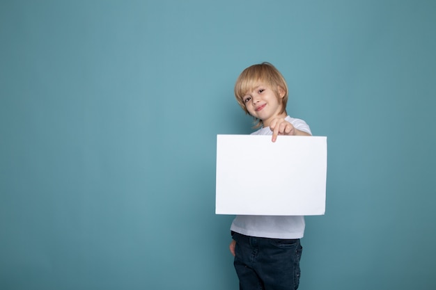 Glimlachende jongen die in witte t-shirt en jeans lege spatie op zijn handen houden tegen covid op blauwe muur