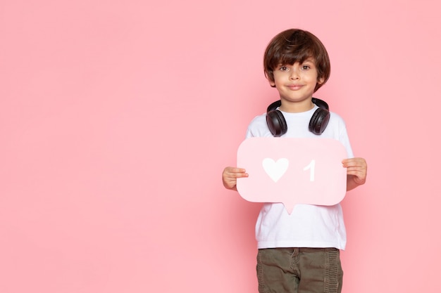 Gratis foto glimlachende jongen in wit t-shirt en kaki broek in zwarte oortelefoons die roze teken met als houden