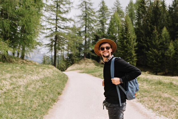 Glimlachende jongen in zwart shirt en hoed poseren op bosweg genieten van reizen in vakantie